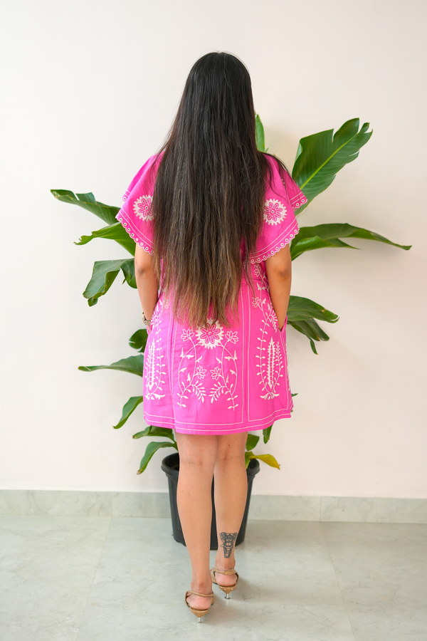 Person wearing a bright pink dress with white patterns, standing in front of a plant against a light background.