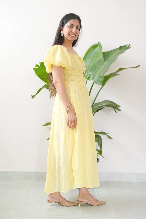 Woman wearing a yellow dress standing in front of a white wall with green plants.