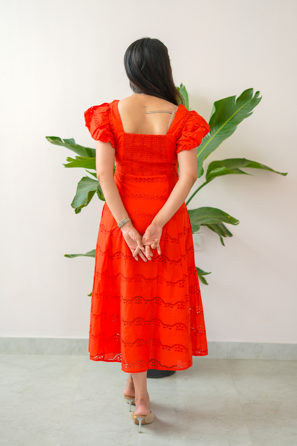 Woman wearing a red dress with puff sleeves standing against a light background with green plants.
