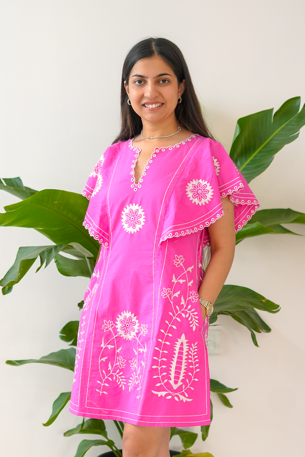 Woman wearing a pink dress with white floral patterns against a white wall with green leaves.