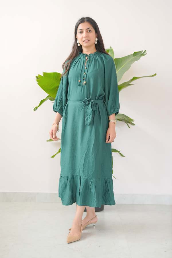 Woman wearing a teal dress standing against a white wall with green leaves.