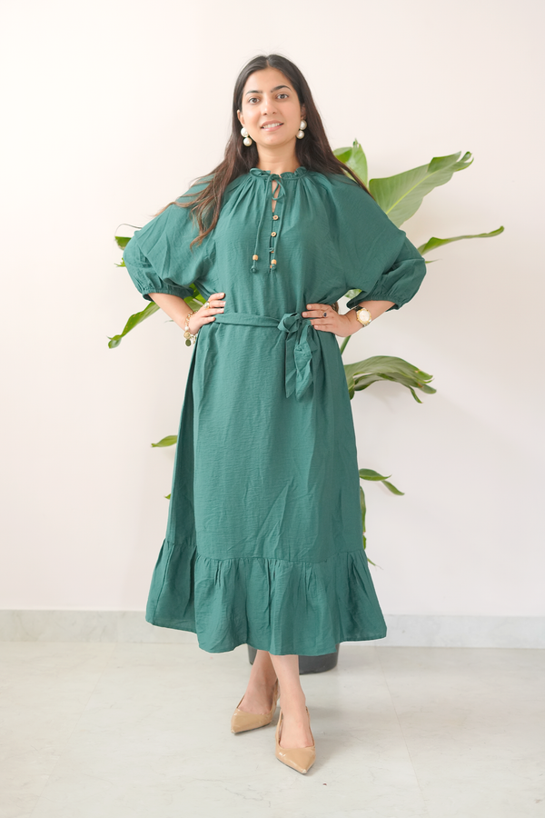 Woman wearing a green dress with puffed sleeves and a belt, standing against a white wall with green leaves.