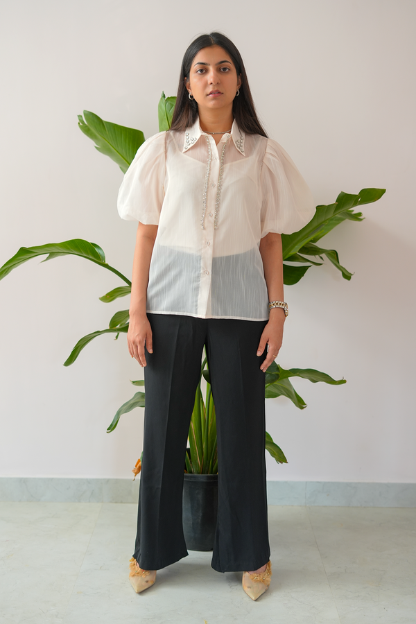 Woman wearing a white blouse with sheer sleeves and black pants standing in front of a plant.