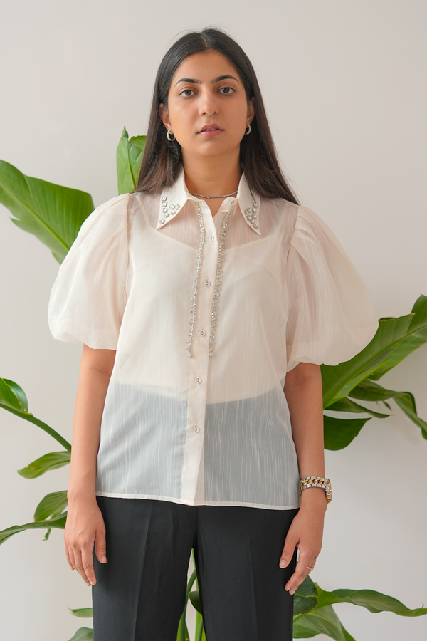 Woman wearing a light-colored blouse with lace details in front of green plants.