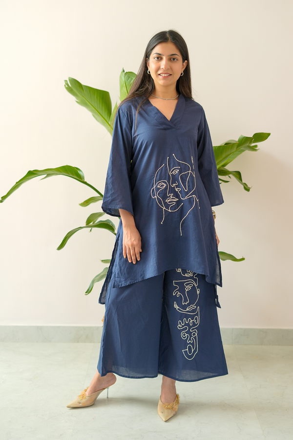 Woman wearing a blue outfit with artistic designs, standing against a light background with plants.