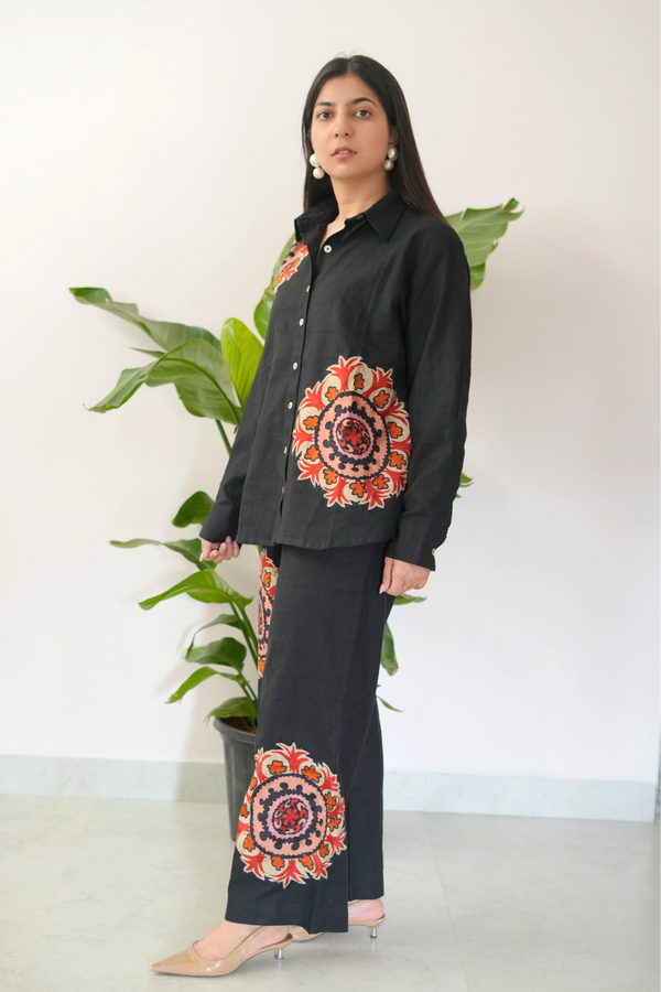 Woman wearing a black outfit with colorful mandala patterns standing in front of a plant.