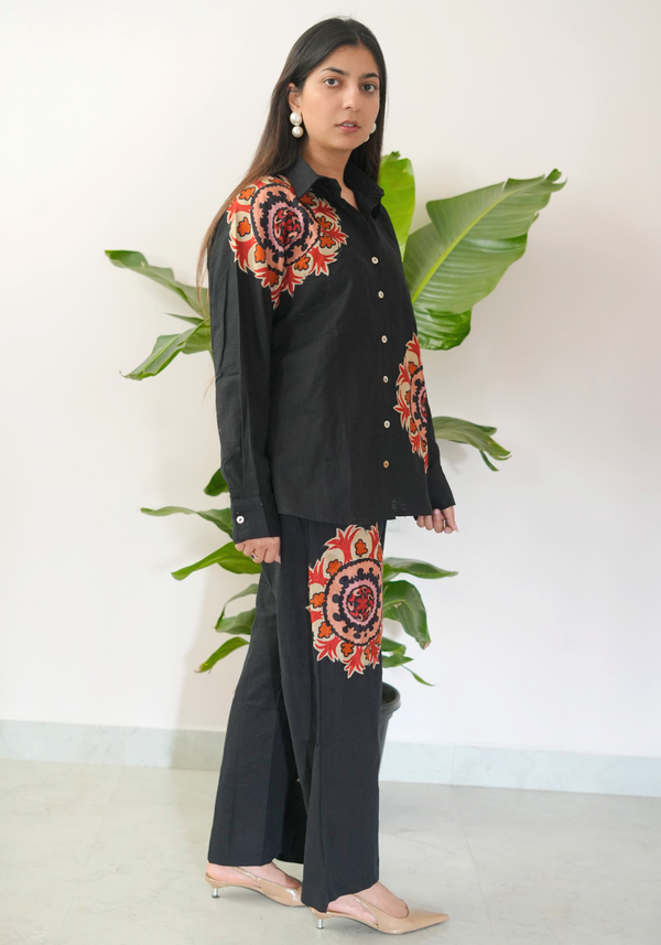 Woman wearing a black outfit with floral embroidery standing against a white background with green plants.
