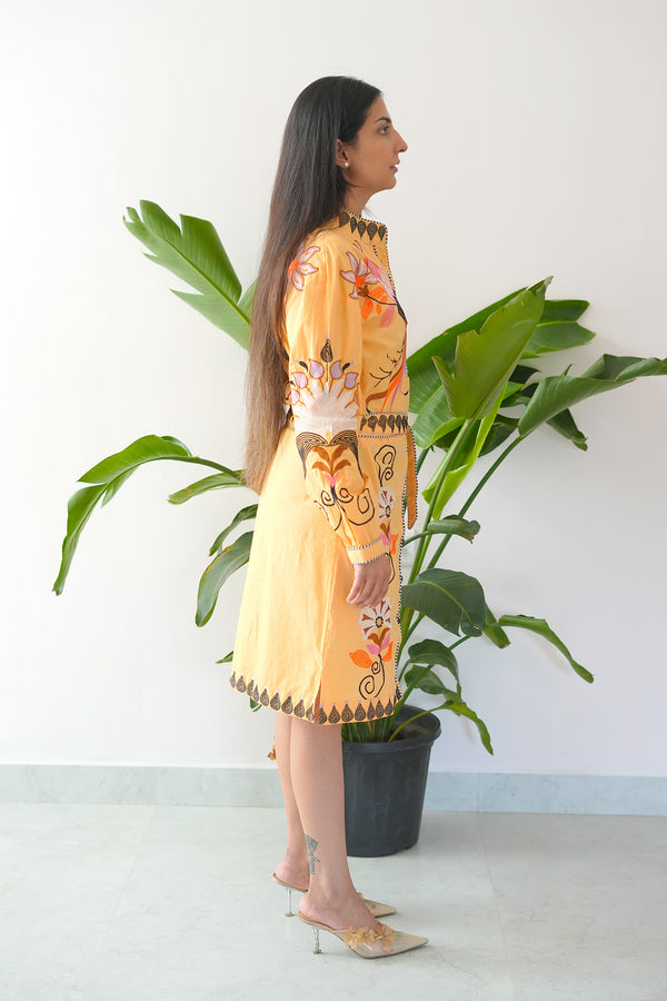 Woman in a yellow floral dress standing next to a potted plant against a white wall.