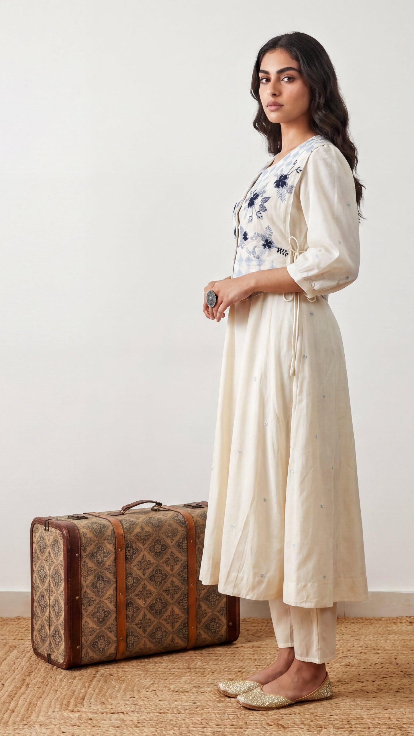 Woman in a white floral dress standing next to a vintage suitcase on a wooden floor.