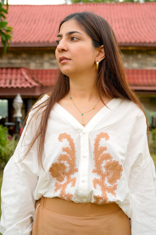 Woman wearing a white blouse with brown embroidery and beige pants outdoors.