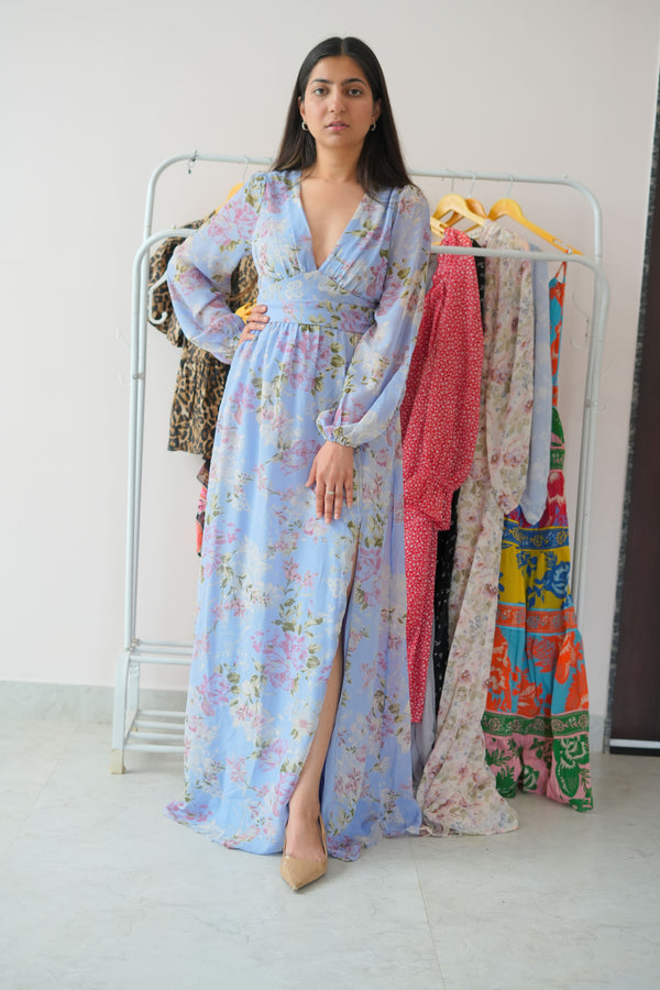 A woman standing in a studio setting wearing a long, floral print, South Asian dress with a V-neck and long sleeves.