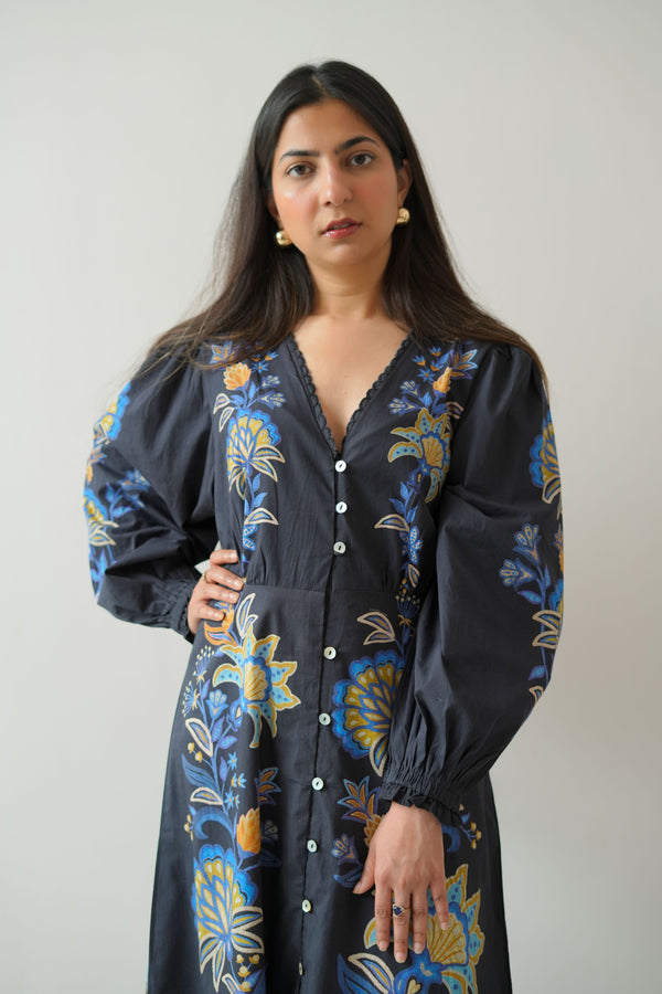 A woman standing against a white background wearing a long navy shirt dress with a floral pattern and button-down front.