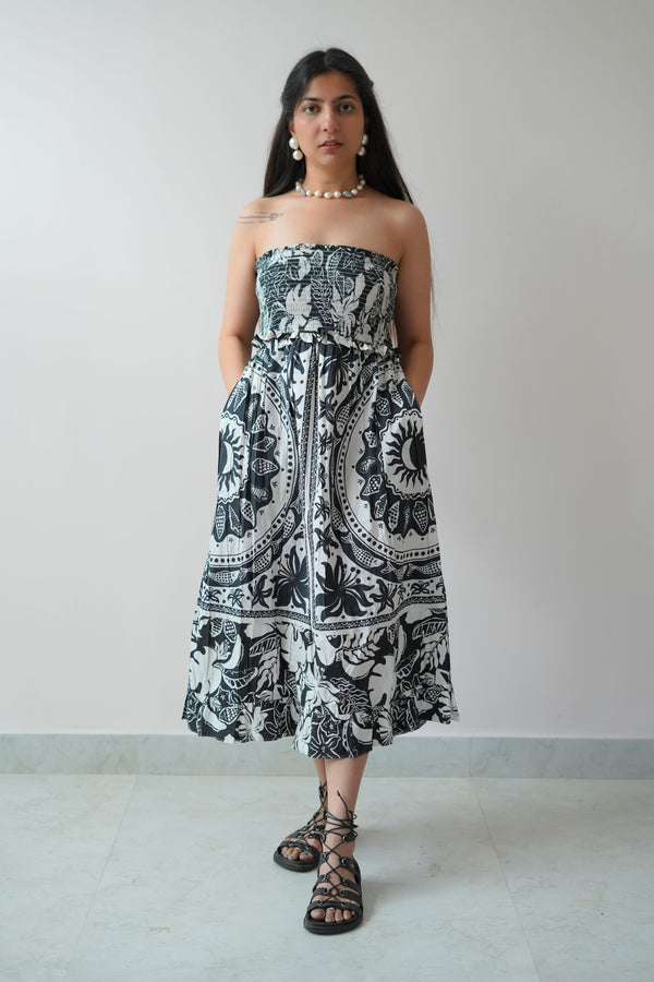 A woman standing in a room with a plain wall, wearing an off-shoulder, long black and white floral dress with pockets, and paired with ankle strap sandals.