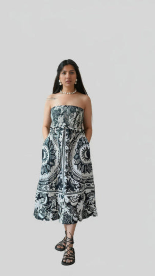 A woman standing in a room with a plain wall, wearing an off-shoulder, long black and white floral dress with pockets, and paired with ankle strap sandals.