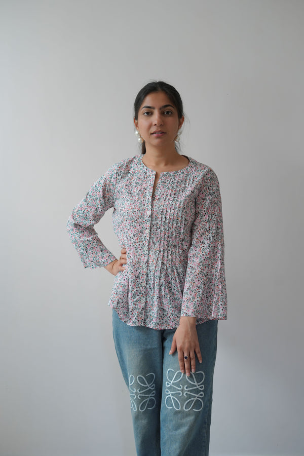 A woman wearing a small flower print top with long sleeves and a round neckline, paired with blue jeans. She is standing against a plain background.