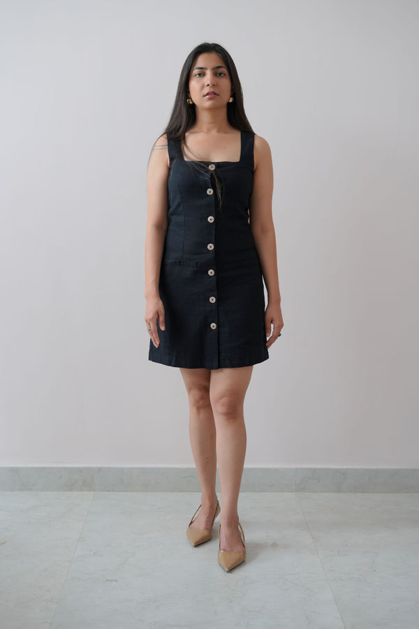 A woman standing against a plain background, wearing a navy blue sleeveless short dress with button-down front.