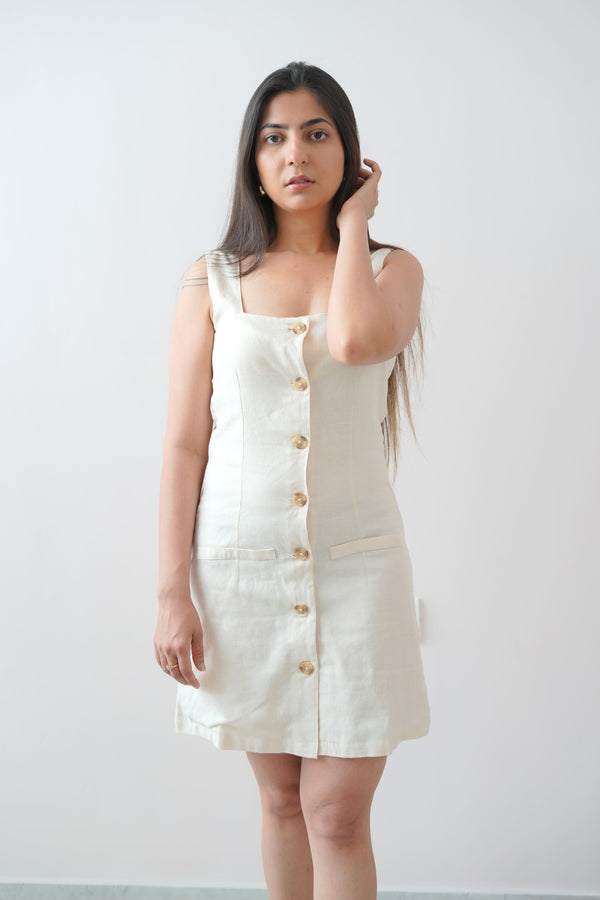 A woman standing in a studio setting, wearing a beige linen short sleeveless dress with square neckline and vertical button details.