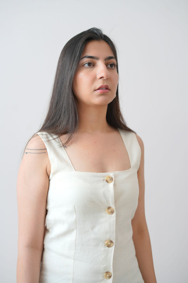 A woman standing in a studio setting, wearing a beige linen short sleeveless dress with square neckline and vertical button details.