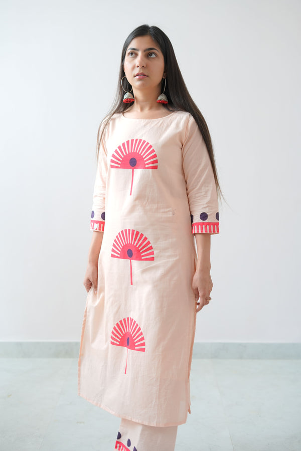 A woman wearing a peach-colored dress with blue geometric patterns standing against a plain background.