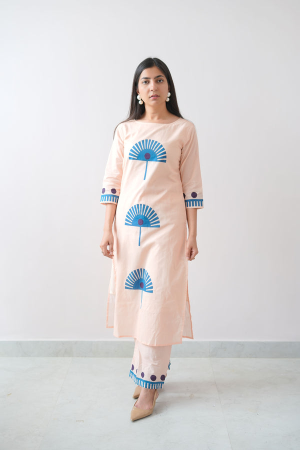 A woman wearing a peach-colored dress with blue geometric patterns standing against a plain background.