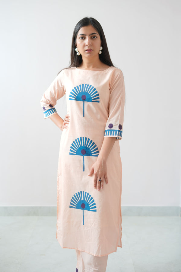 A woman wearing a peach-colored dress with blue geometric patterns standing against a plain background.