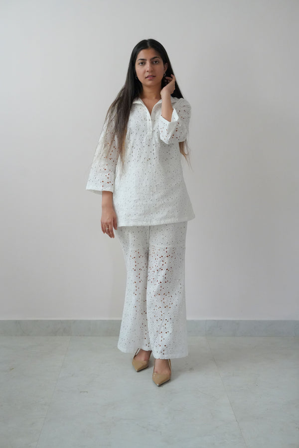 A woman wearing a white long-sleeve top with a classic collar and cutwork detailing, paired with matching white straight-leg pants.