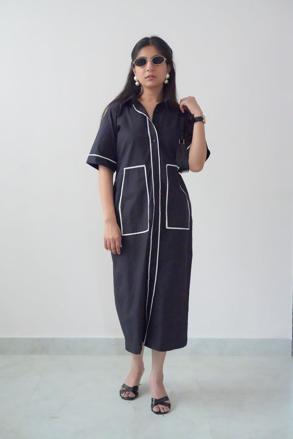 A person wearing a navy dress with white button-down front and oversized pockets, paired with sunglasses and open-toe heeled sandals.