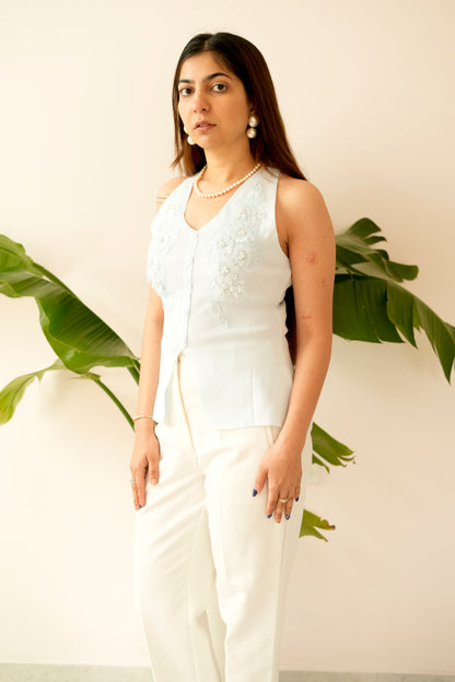 Woman wearing a white sleeveless top and pants standing against a light background with green leaves.