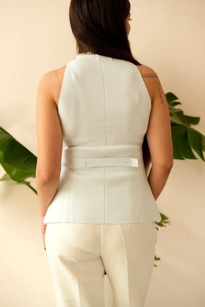 Person wearing a white sleeveless top and beige pants against a neutral background with greenery.