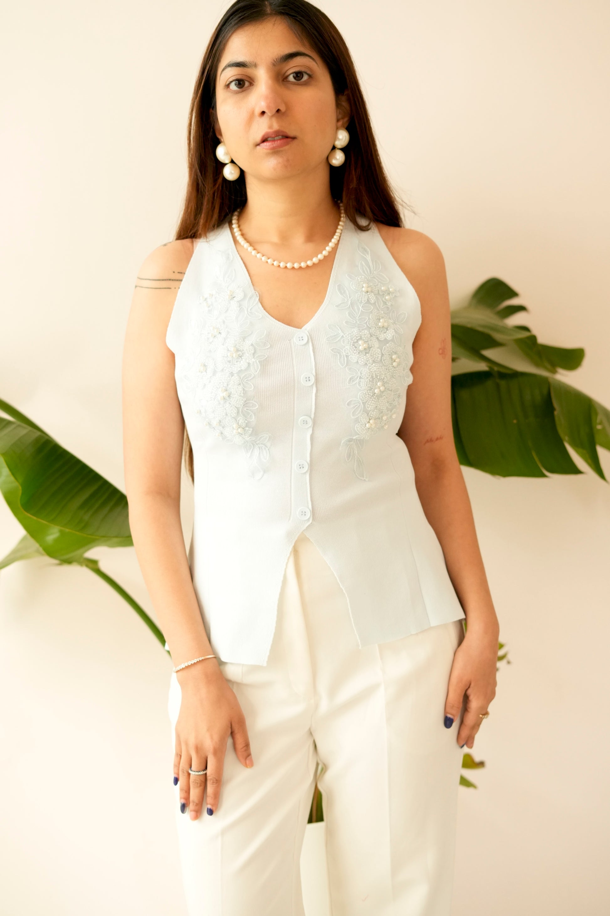 Woman wearing a white sleeveless top with floral patterns and white pants, standing against a light background with green leaves.