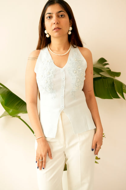 Woman wearing a white sleeveless top with floral patterns and white pants, standing against a light background with green leaves.