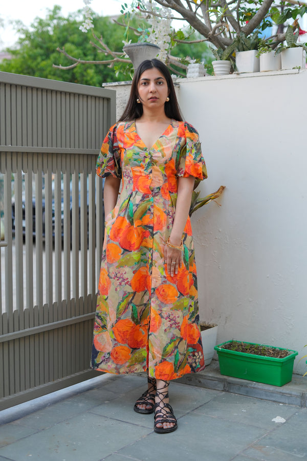 A woman standing in an outdoor setting wearing a vibrant orange long bodycon dress with floral prints, a V-neckline, short sleeves, and side pockets.
