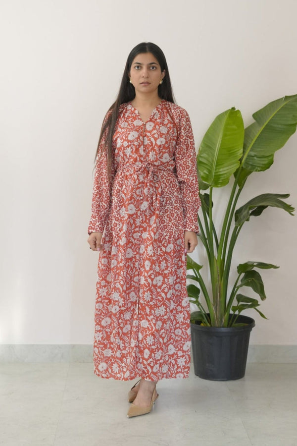 A woman standing in front of a potted plant, wearing a brick red maxi dress with white floral patterns, long sleeves, and a tied waist.