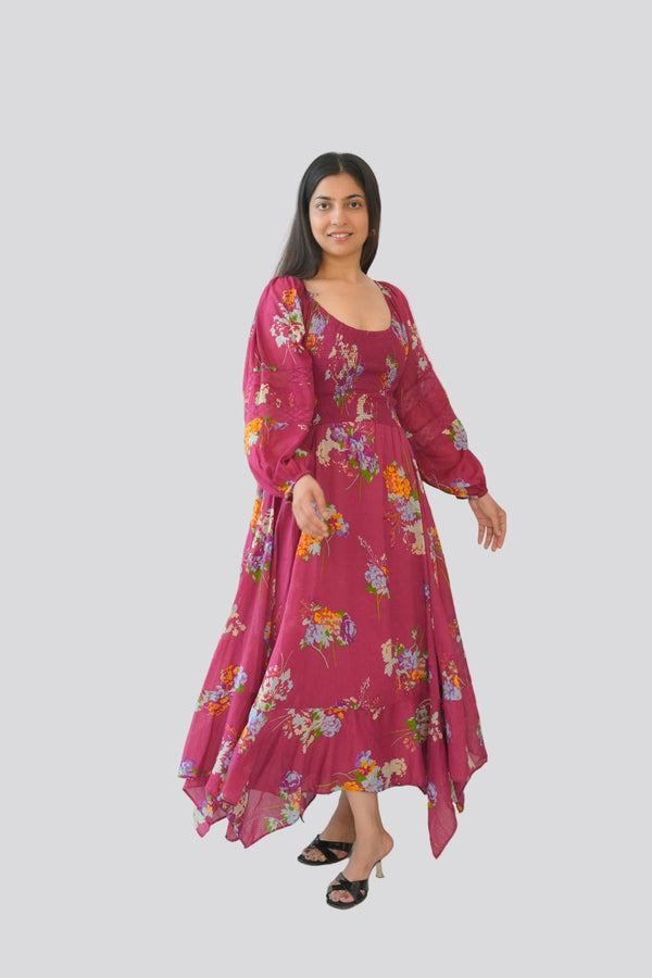 Woman in a pink floral dress standing next to a large green plant against a white wall.