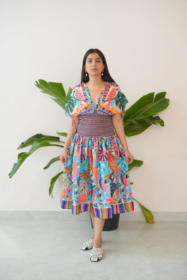 Woman in a colorful dress standing next to a potted plant against a white wall.