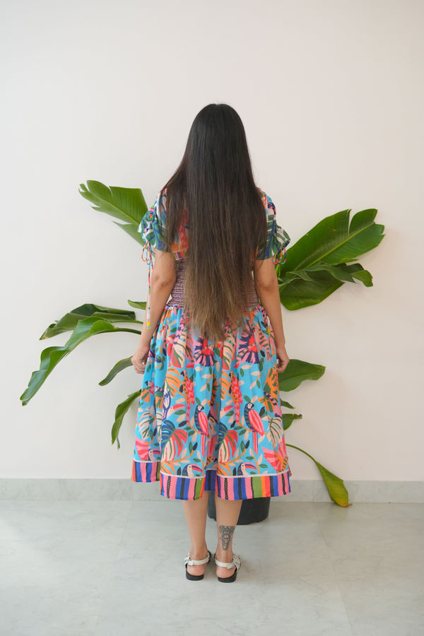 Person wearing a colorful dress standing among potted plants on a white wall.