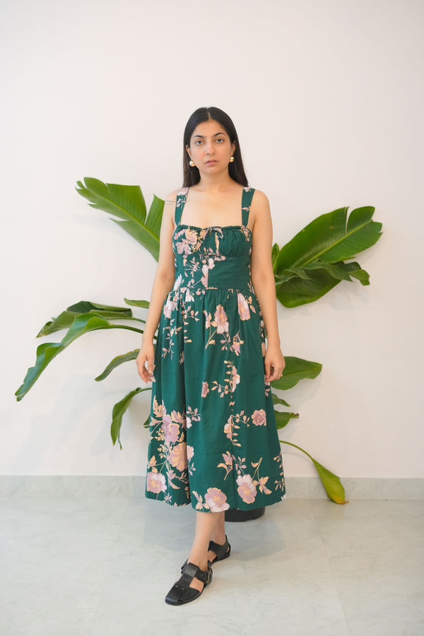 Woman wearing a green floral dress standing against a white wall with large green leaves.