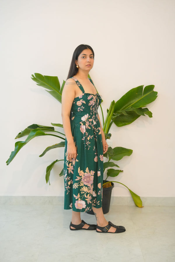 Woman in a green floral dress standing next to a large plant indoors.