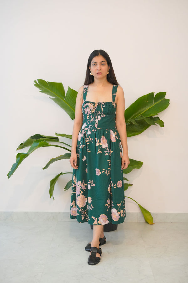 Woman in a floral dress standing next to a large plant against a white wall