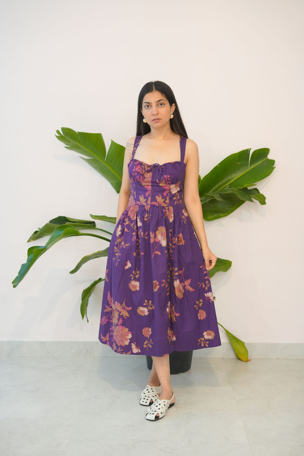 Woman in a purple floral dress standing next to a large green leafy plant on a white background