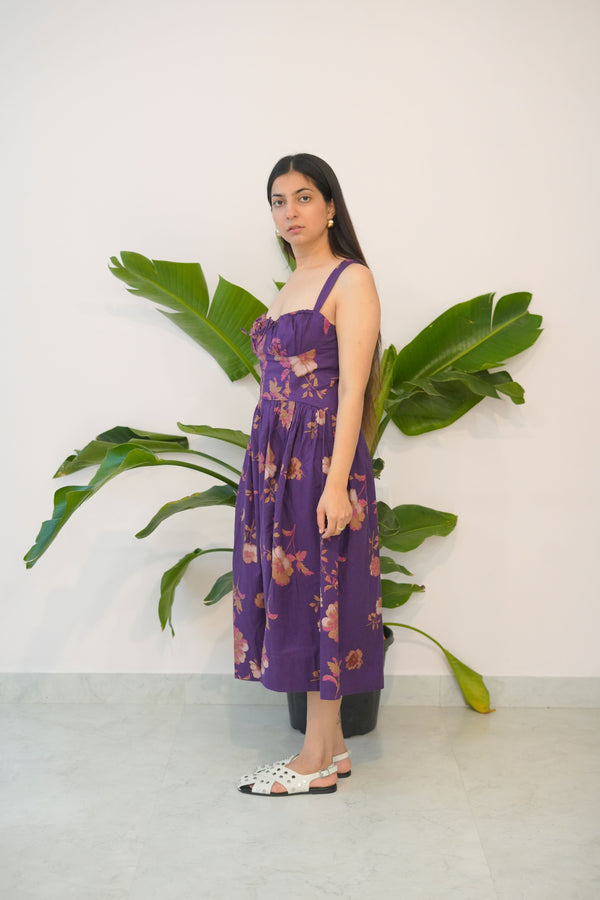 Woman in a purple dress standing next to a large green plant against a white wall.