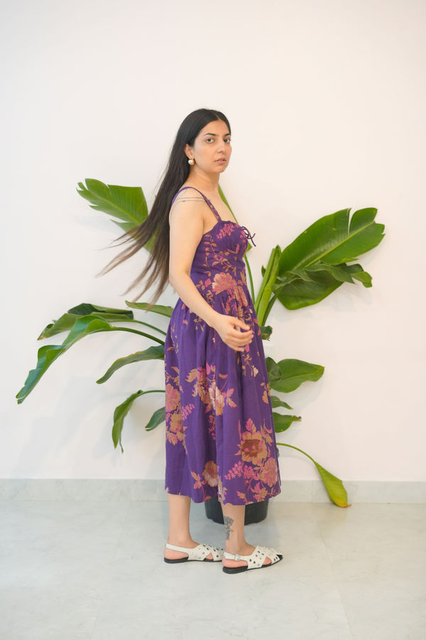 Woman in a purple dress standing next to a large green plant against a white wall.