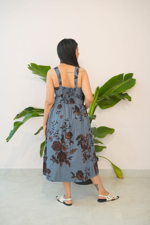 Person wearing a blue floral dress standing next to a large green leafy plant indoors.