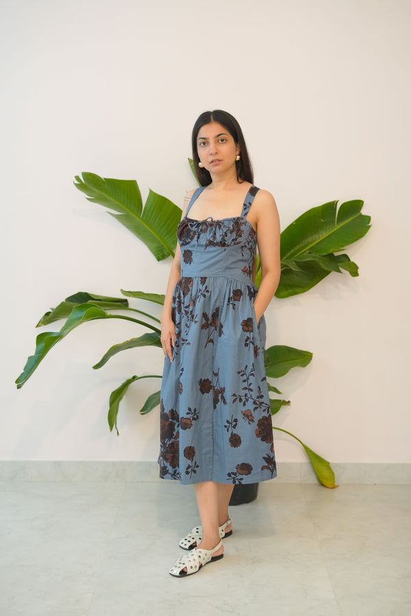 Woman in a blue floral dress standing next to a large green plant indoors.