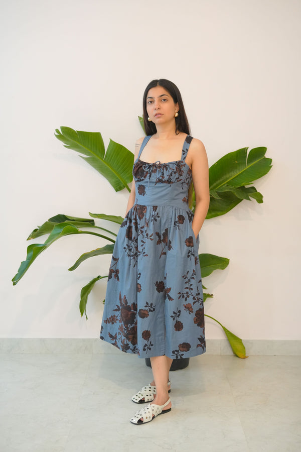 Woman in a blue floral dress standing next to a large green plant indoors.