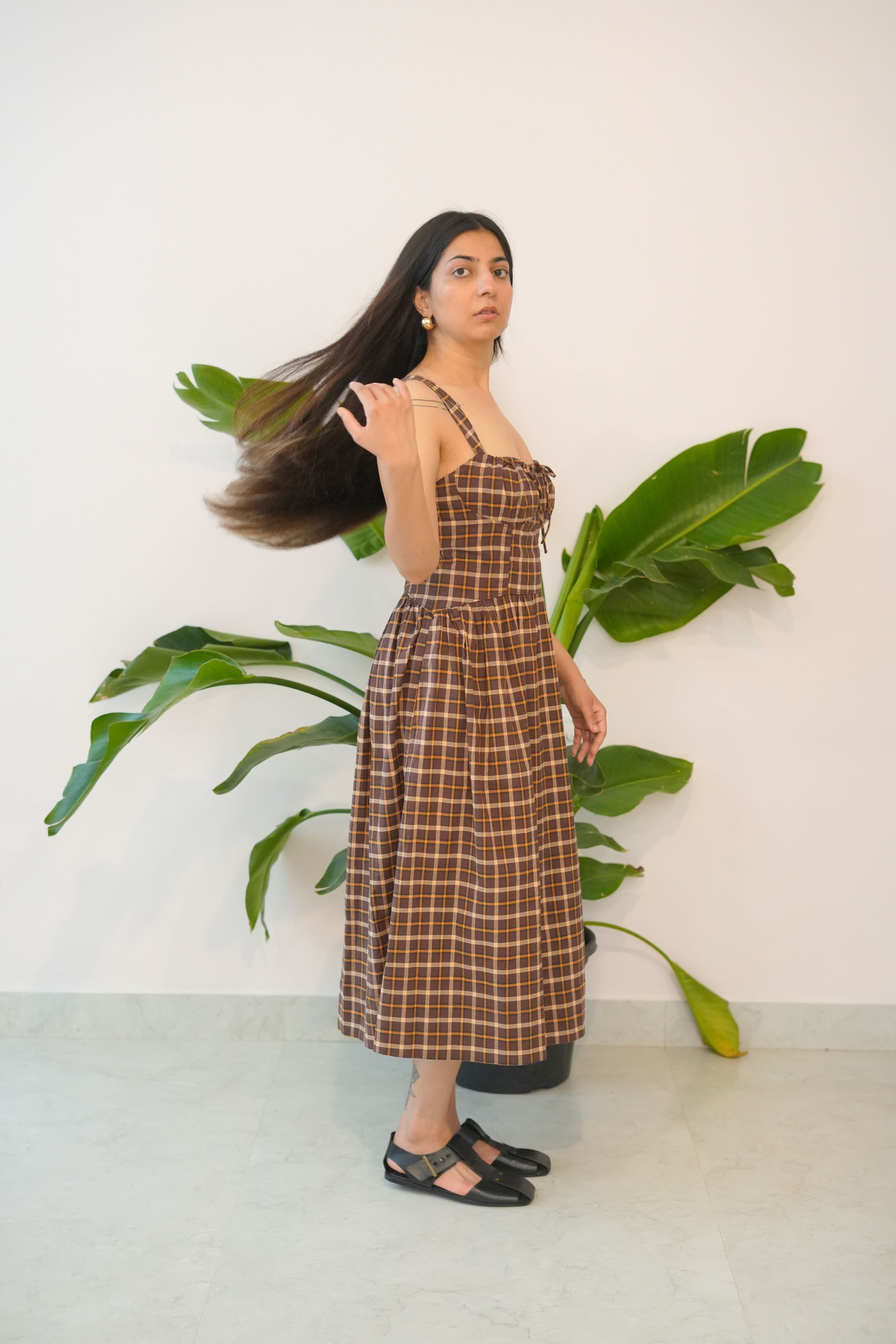Woman in a plaid dress standing next to a plant against a white wall