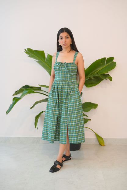 Woman in a green checkered dress standing between large green leaves against a white wall.
