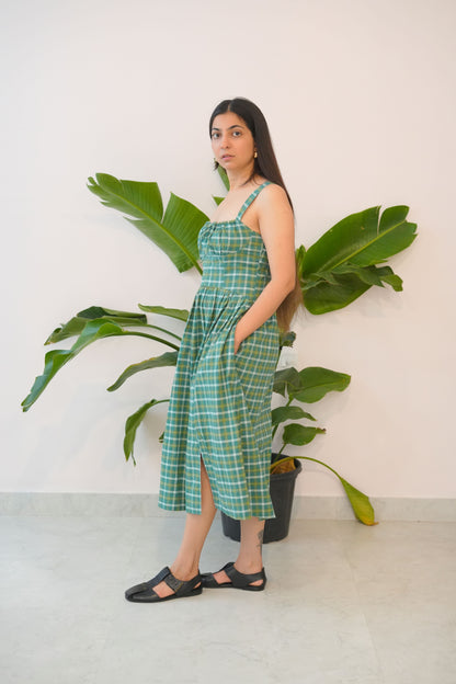 Woman in a green checkered dress standing next to a large plant against a white wall.