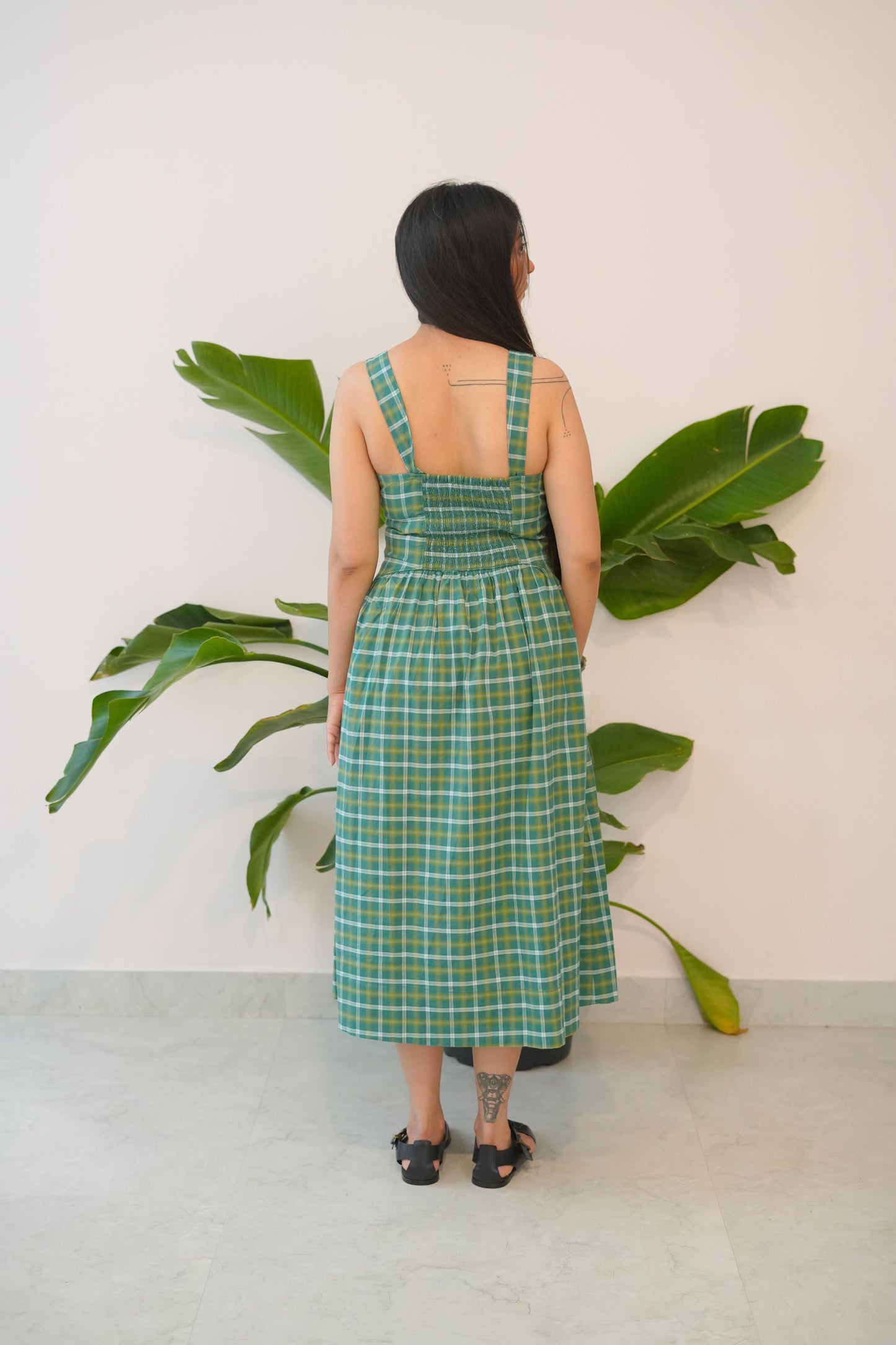 Person wearing a green plaid dress standing between two large plants indoors.