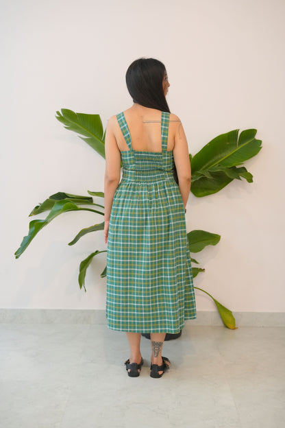 Person wearing a green plaid dress standing between two large plants indoors.